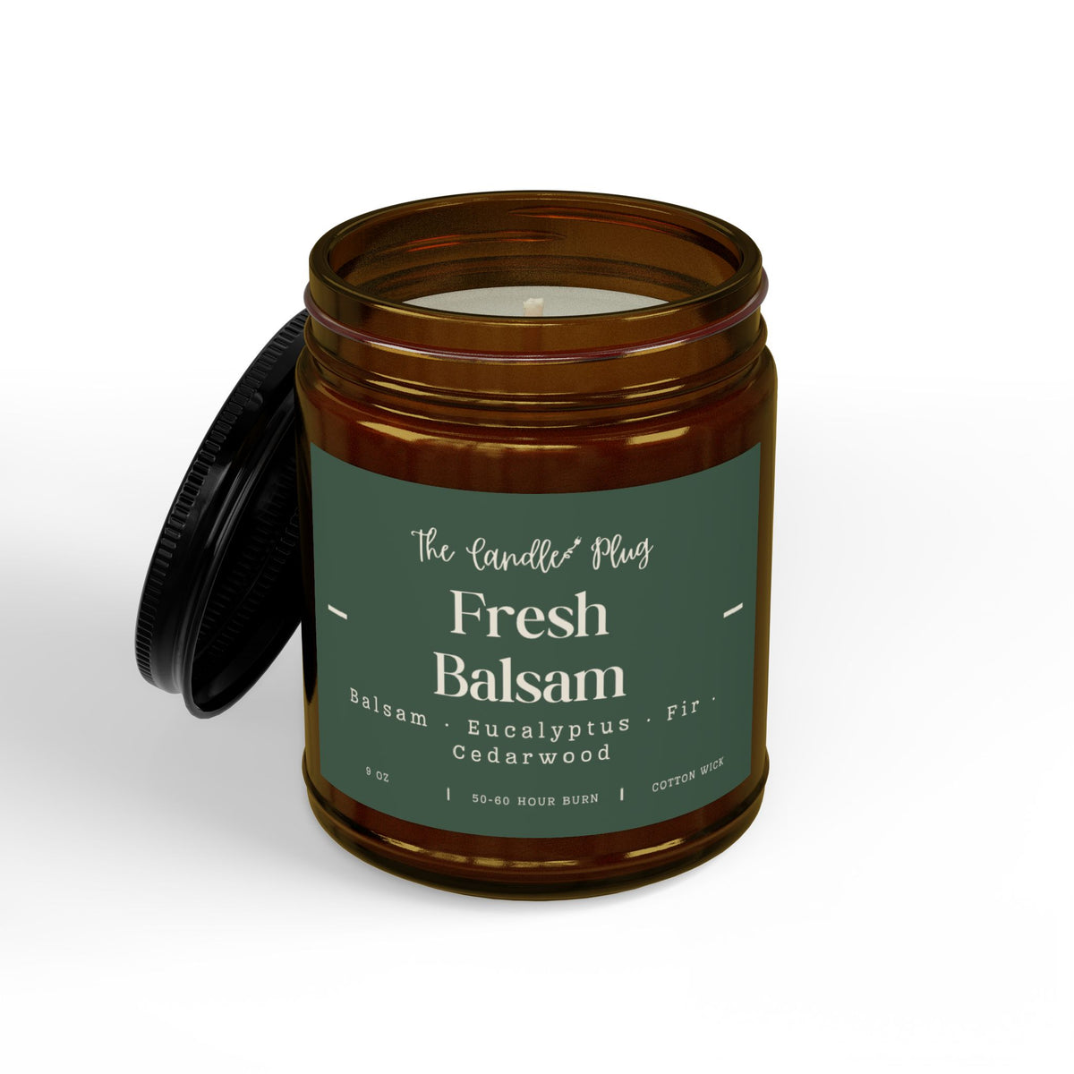 An amber glass jar candle with its black lid leaning against it on a clean white background. The candle features a dark green label with white text that reads 'The Candle Co Fresh Balsam' and lists notes of cedarwood, eucalyptus, and fir.