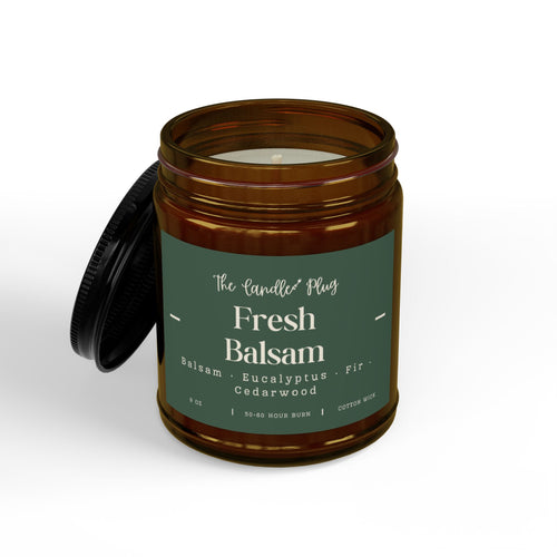 An amber glass jar candle with its black lid leaning against it on a clean white background. The candle features a dark green label with white text that reads 'The Candle Co Fresh Balsam' and lists notes of cedarwood, eucalyptus, and fir.